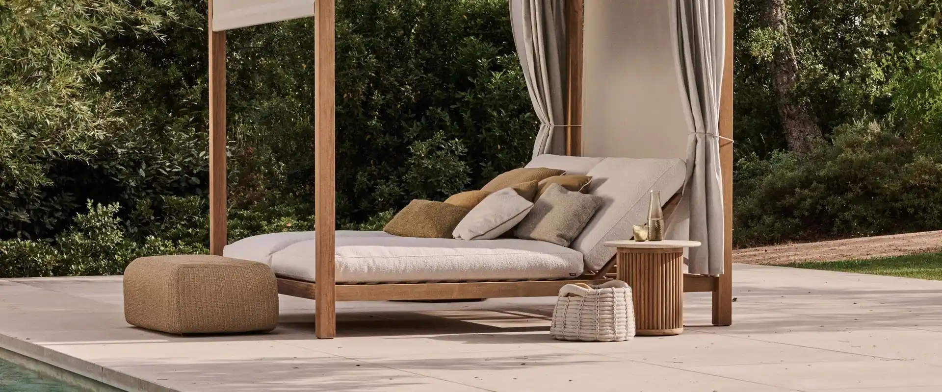 Tribù teak canopy daybed with white curtains and cushions beside a pool, with poufs and green woods behind