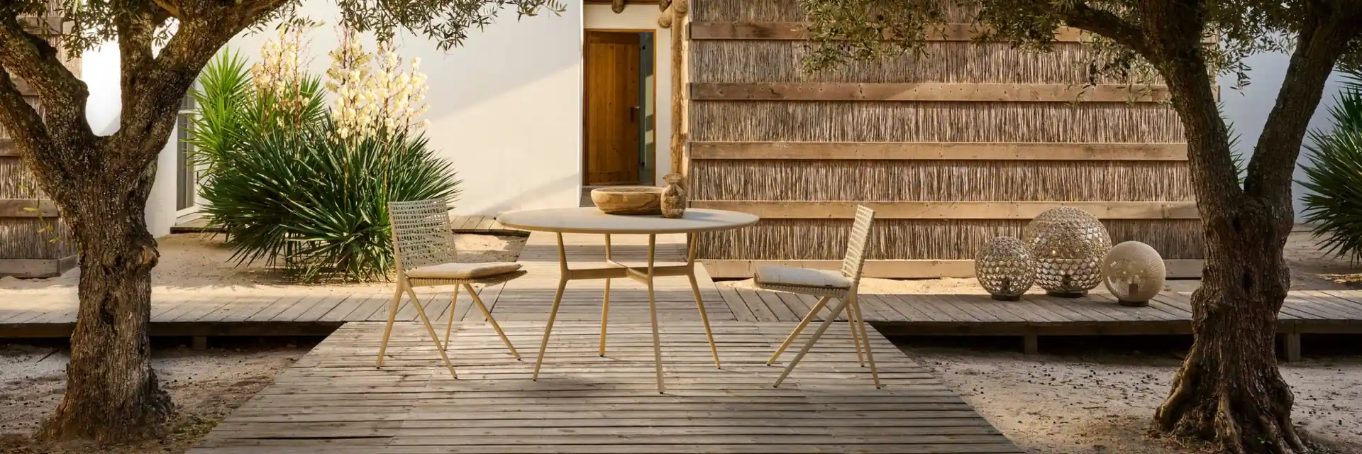 Tribù dining chairs in yellow around a small table on a wooden deck between two old olive trees, a reed-clad house behind