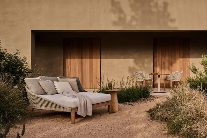 Tribù double daybed with cushions and a throw in front of a rammed-earth house with timber shutters, among ornamental grasses