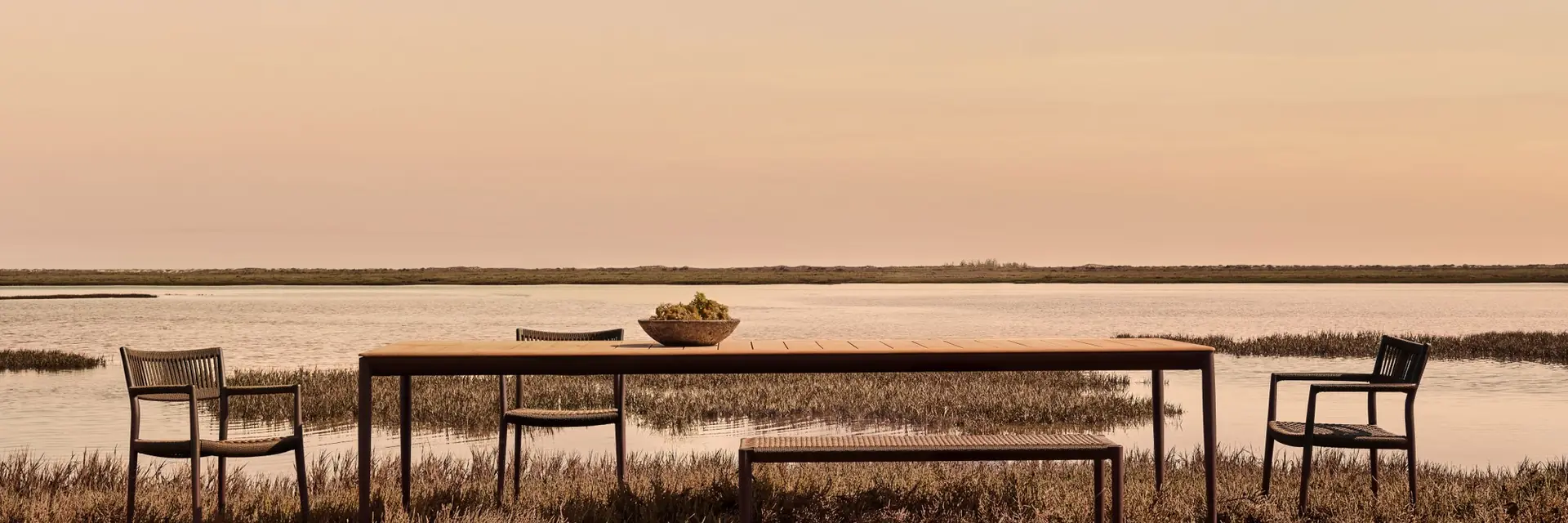 Tribù dining table with a bench and armchairs on a marshy shoreline at dusk