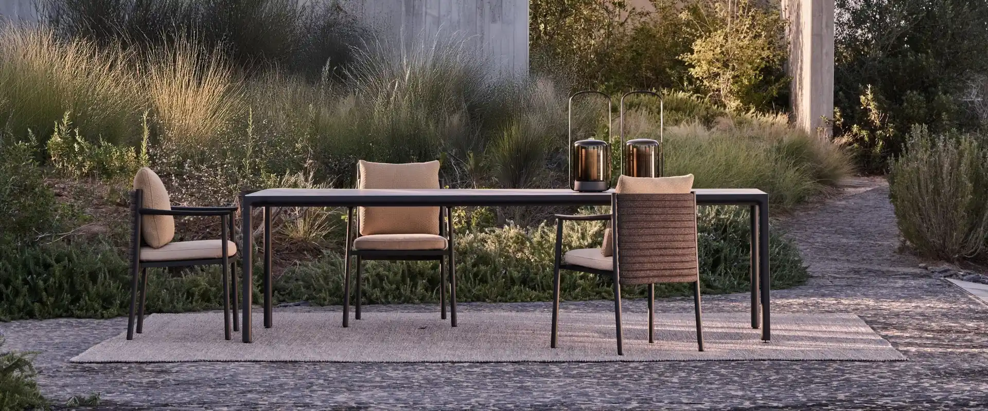 Tribù long dining table with woven chairs on a gravel terrace beside a concrete house and tall grasses