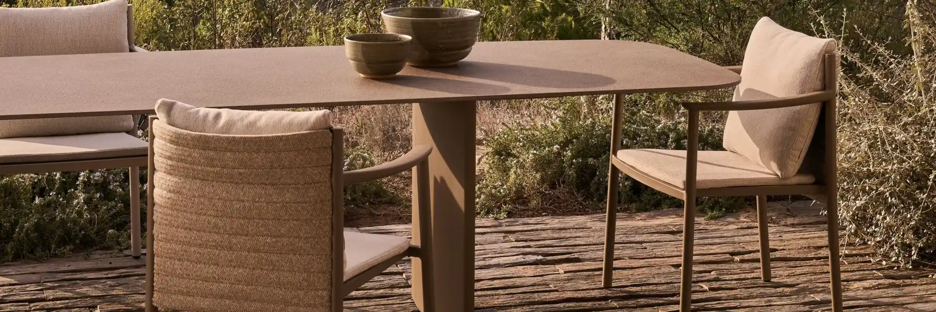 Tribù dining table with woven armchairs on a timber deck, with shrubs behind
