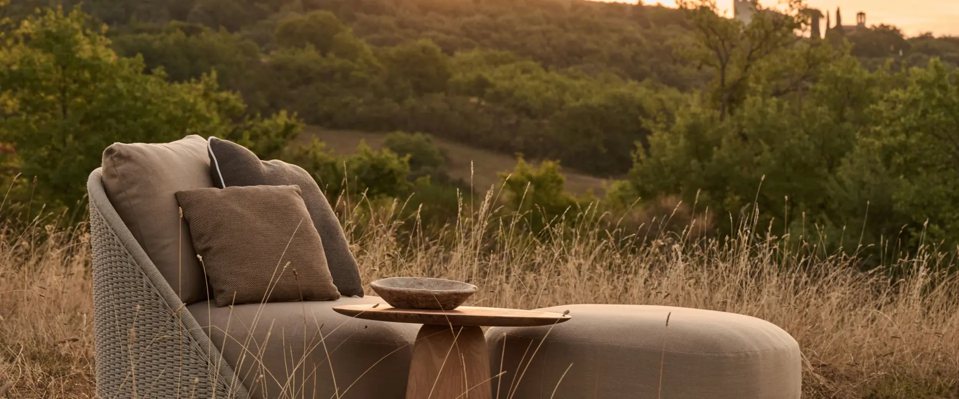 Tribù Dunes teak side table beside a round daybed on a path through a meadow at sunset