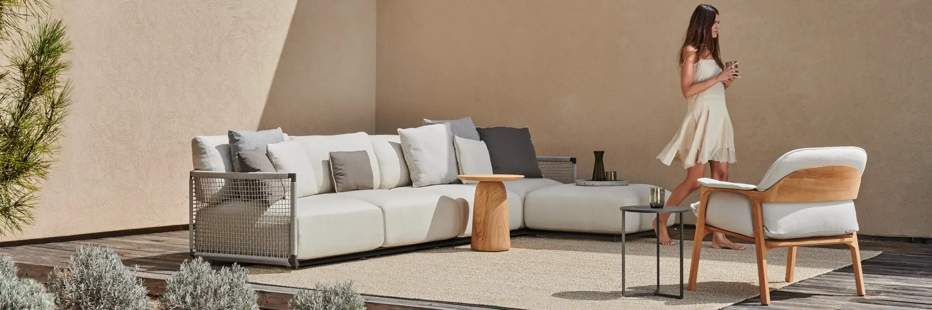 Tribù Dunes modular sofa and an armchair on a timber deck beside a sand-coloured wall, with a woman walking past