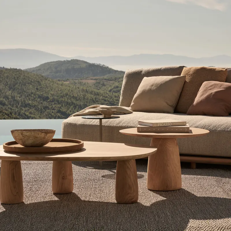 Tribù Dunes low table and side table in teak beside an outdoor sofa on a terrace overlooking wooded hills