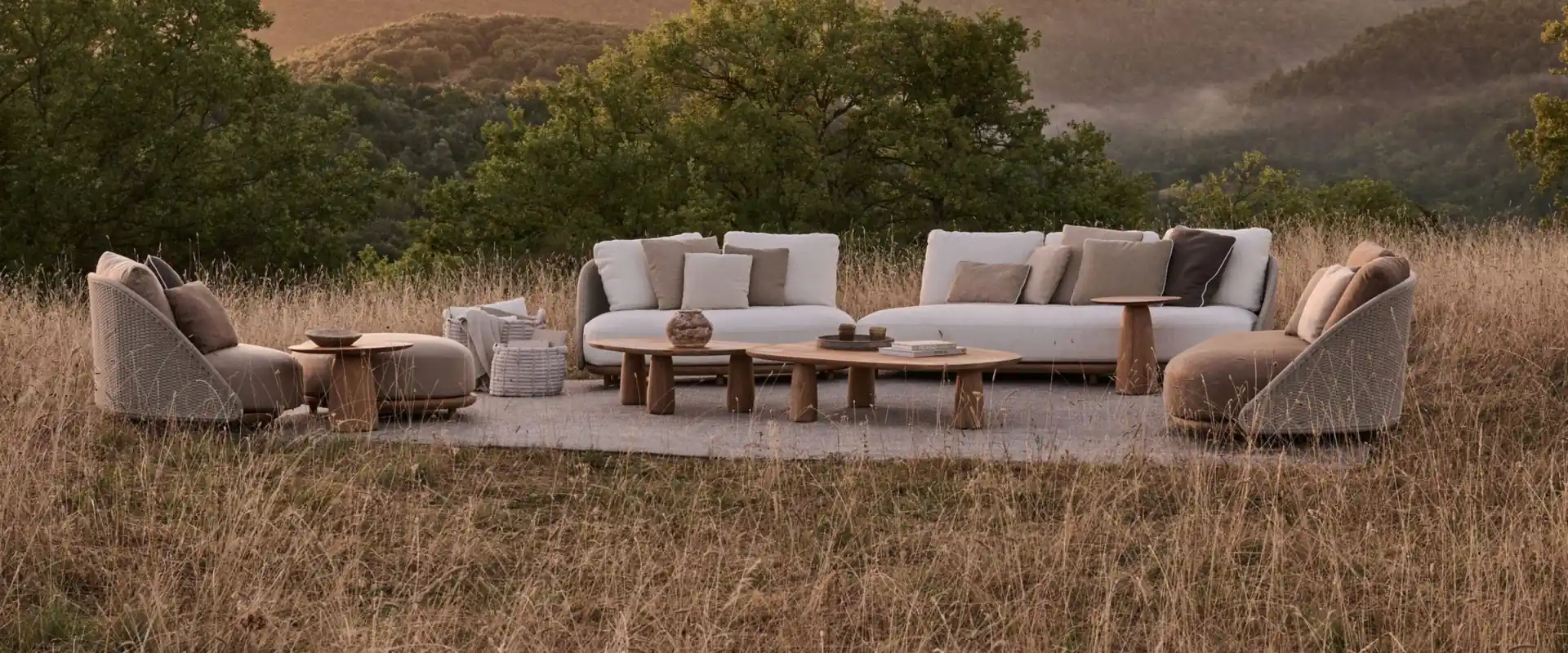 Tribù Elio sofas and woven armchairs around low tables in a golden meadow, with wooded hills behind at sunset
