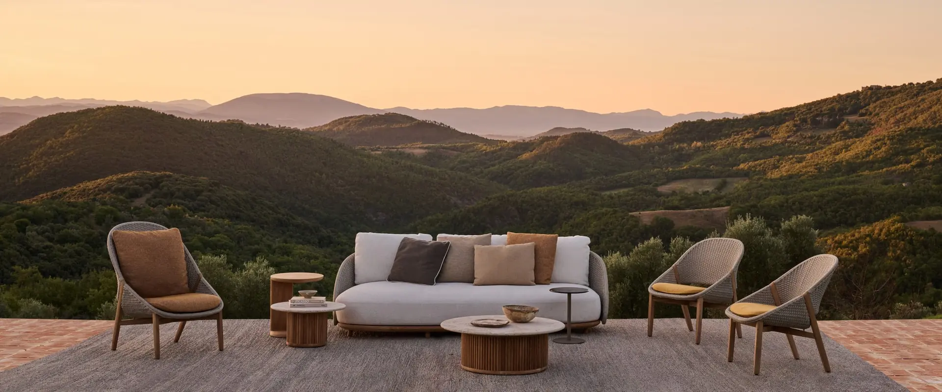 Tribù Elio sofa and woven lounge chairs on a brick terrace at sunset, overlooking wooded hills