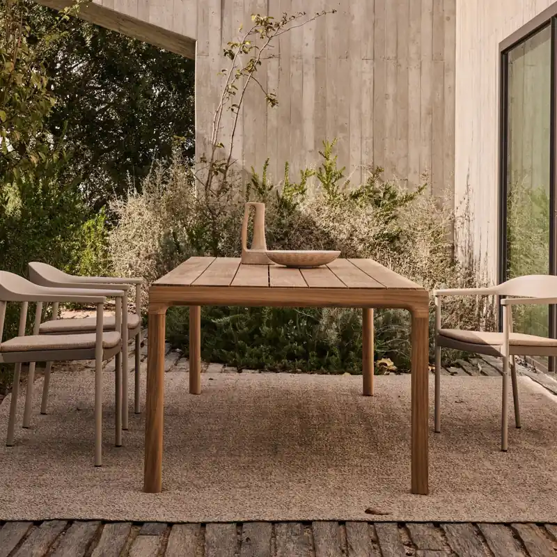 Tribù Illum teak dining table with four chairs on a rug beside a concrete house and wild grasses