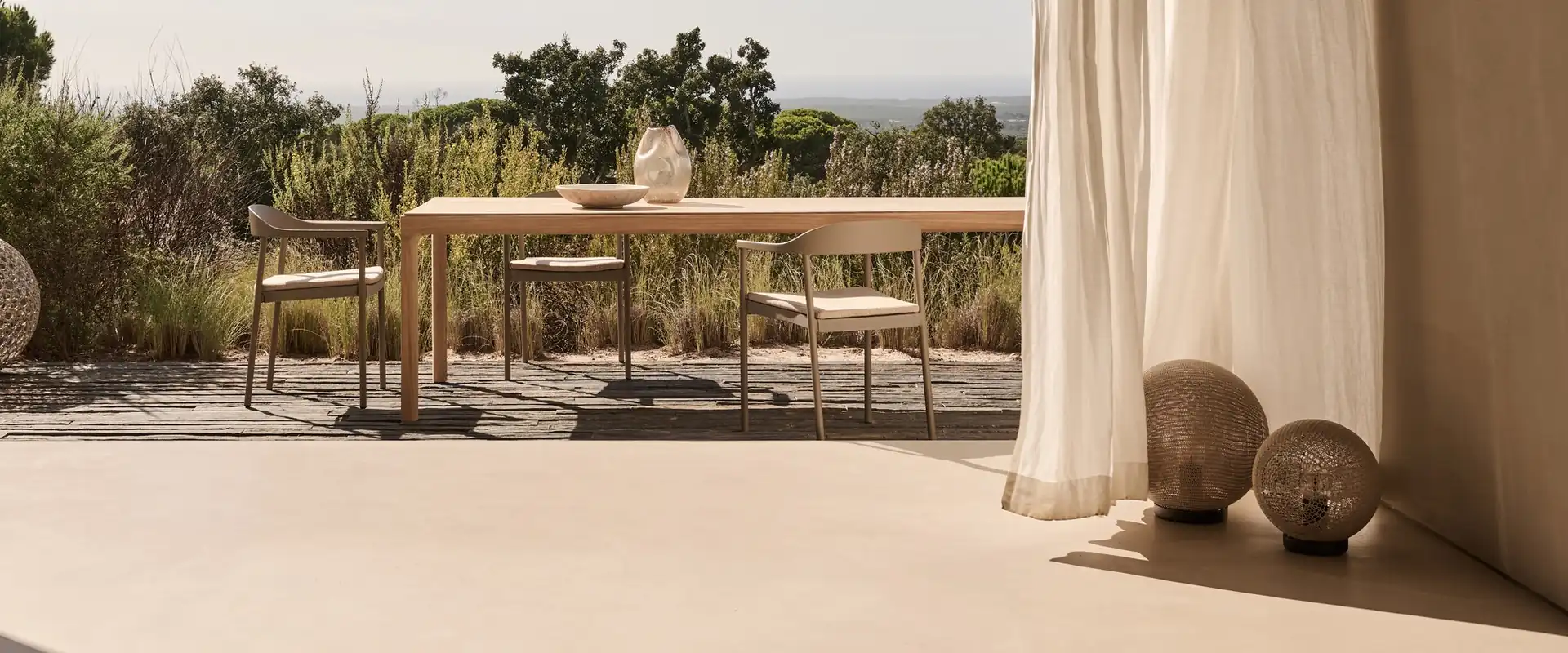 Tribù Illum dining table and chairs beneath a gabled porch with linen curtains, looking out over scrubland