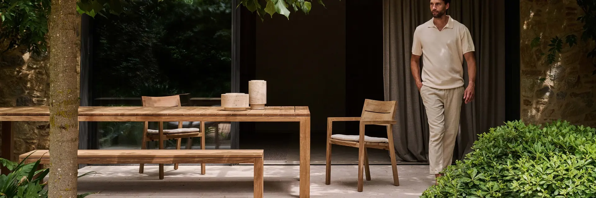 Tribù Kos teak dining table, bench and armchairs under a tree beside a dark pavilion, with a man standing by