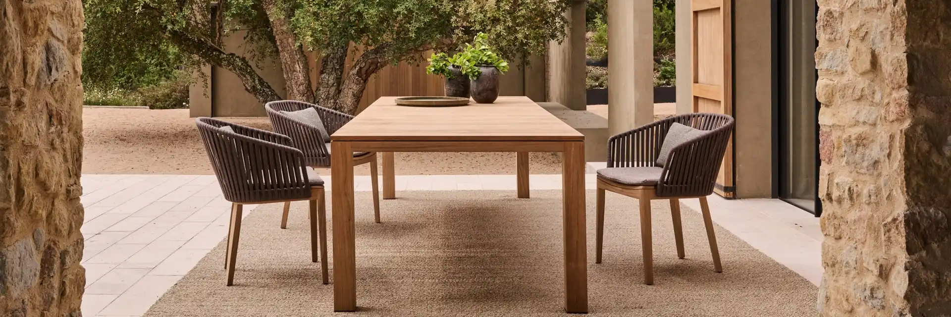 Tribù Kos teak dining table and chairs on a terrace between stone columns, with olive trees behind