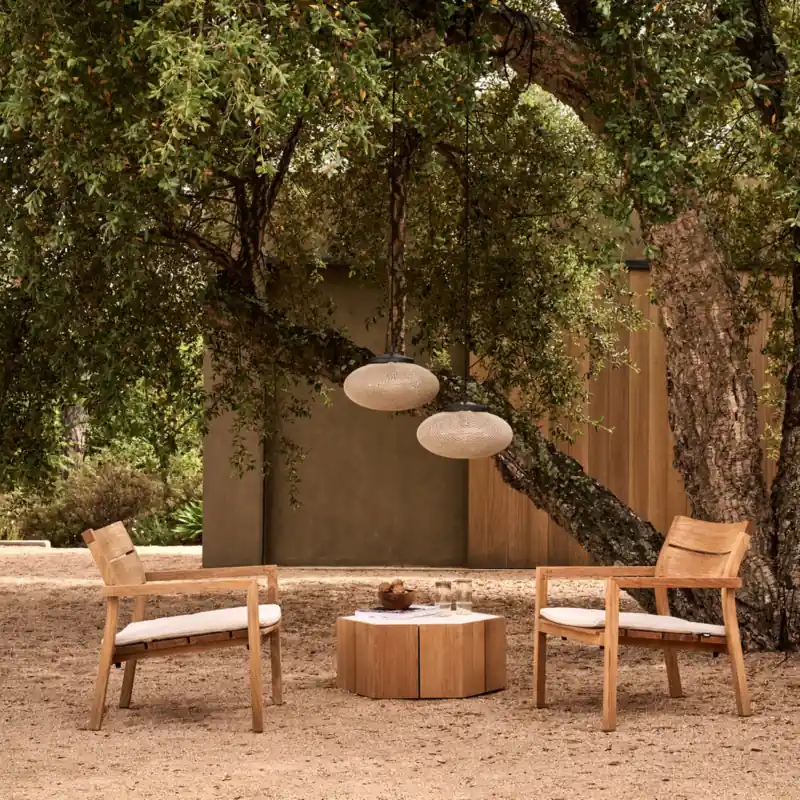 Two Tribù Kos teak lounge chairs with a round side table on gravel under an old tree hung with lamps