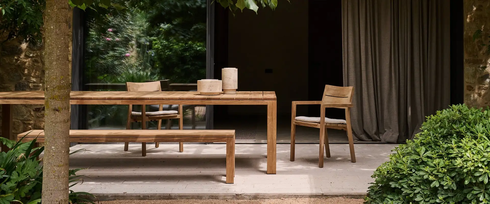 Tribù Kos teak dining table with a bench and an armchair under a tree, before a house with dark curtains