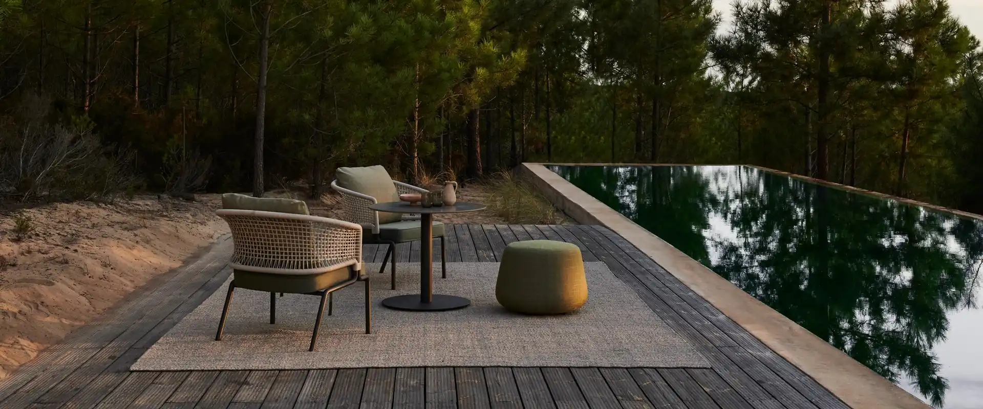 Tribù woven lounge chair with a side table and a green pouf on a wooden deck beside a dark pool, among pine trees