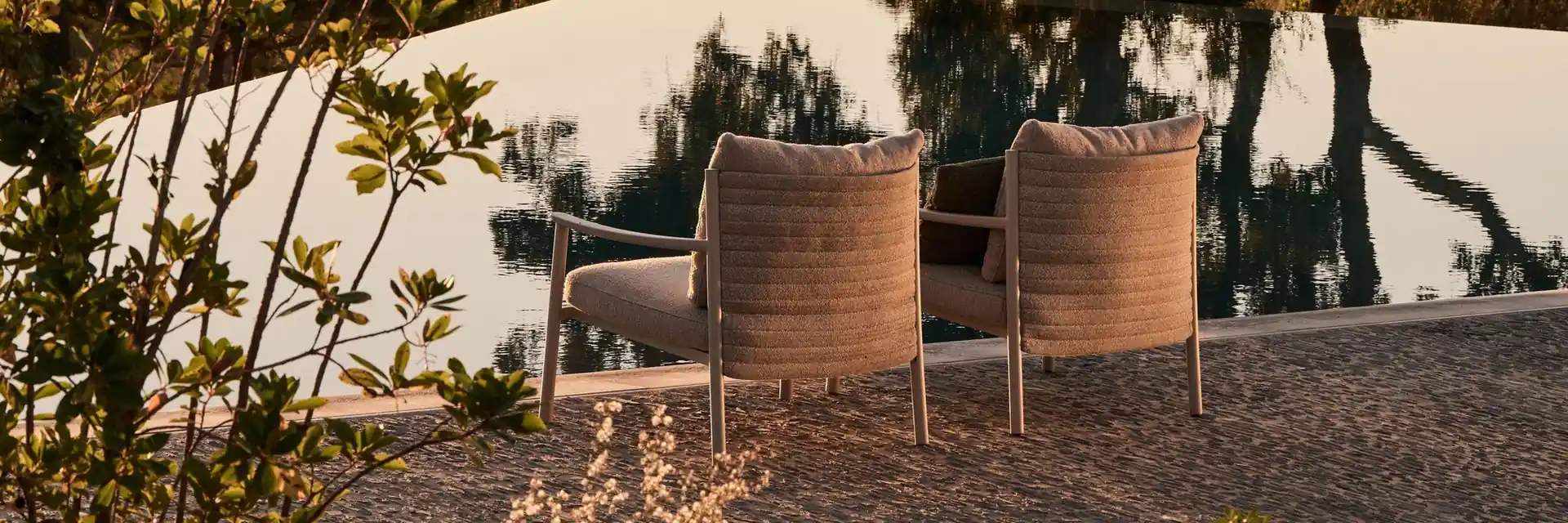 Two Tribù woven lounge chairs at the edge of a still pool that mirrors the trees, in warm evening light