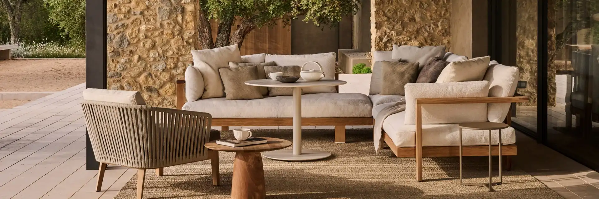 Tribù sofas, armchairs and side tables on a rug under the roof of a covered terrace with stone walls