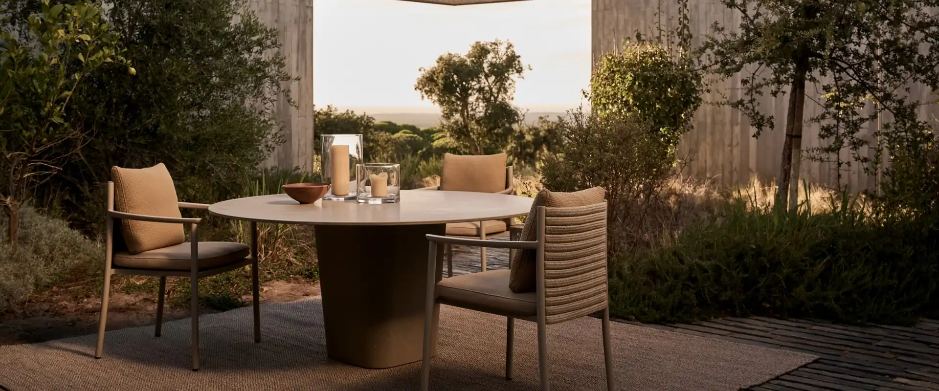 Tribù Lucca dining armchairs with tan cushions around a round table in a concrete courtyard with wild plants