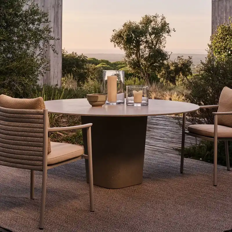 Two Tribù Lucca dining armchairs with woven backs at a round table set with glasses, on a rug among shrubs