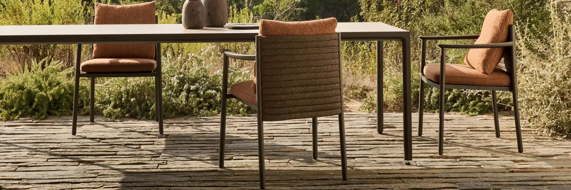 Tribù Lucca dining armchairs with orange cushions at a slim dark table on a deck, overlooking scrubland