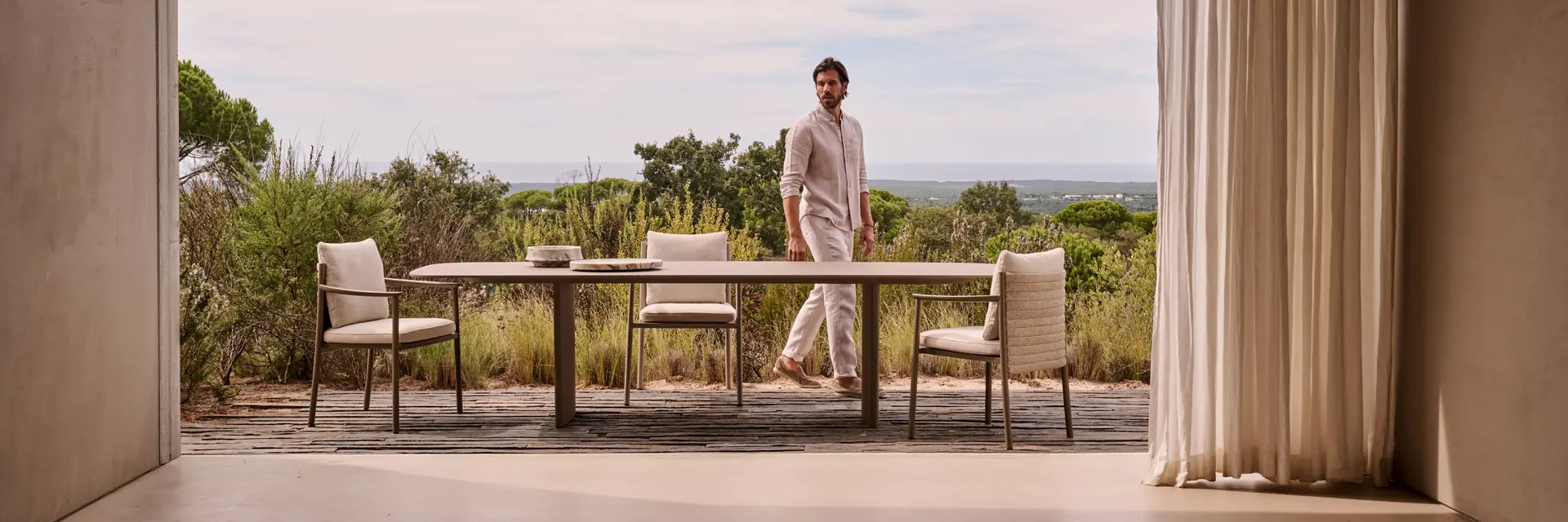 Tribù Lucca dining table and chairs on a terrace seen from under a pitched roof, with a man standing by and scrubland beyond at sunset
