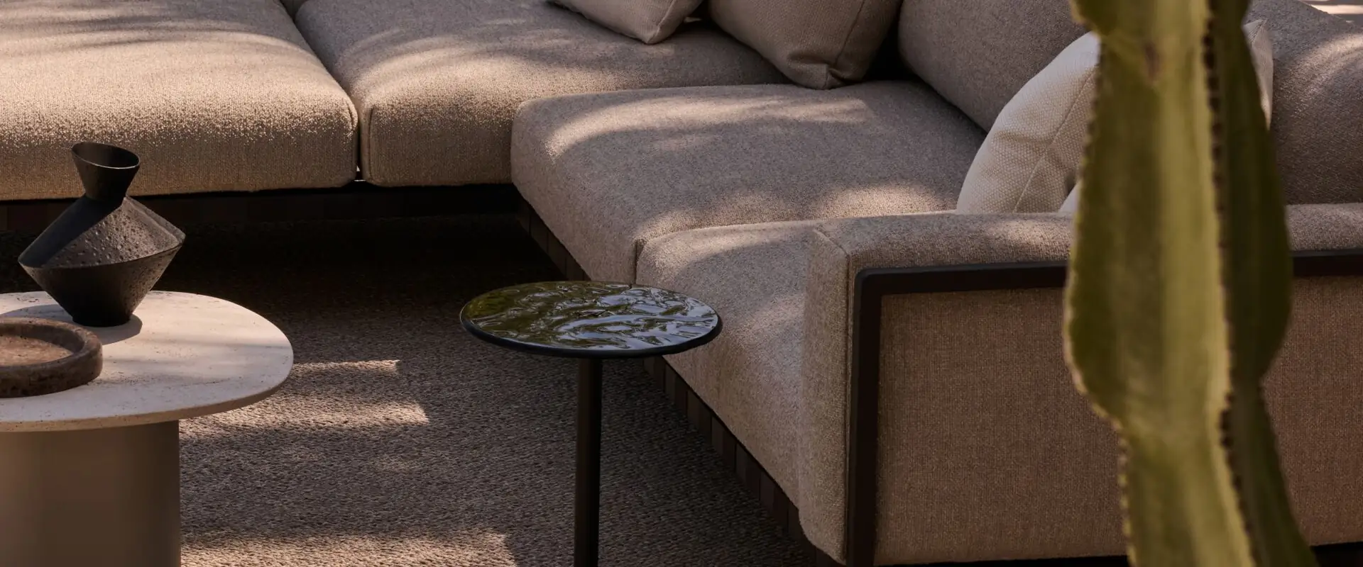 Tribù Mare side table with a dark glass top beside a corner sofa on a terrace, with a tall cactus in front