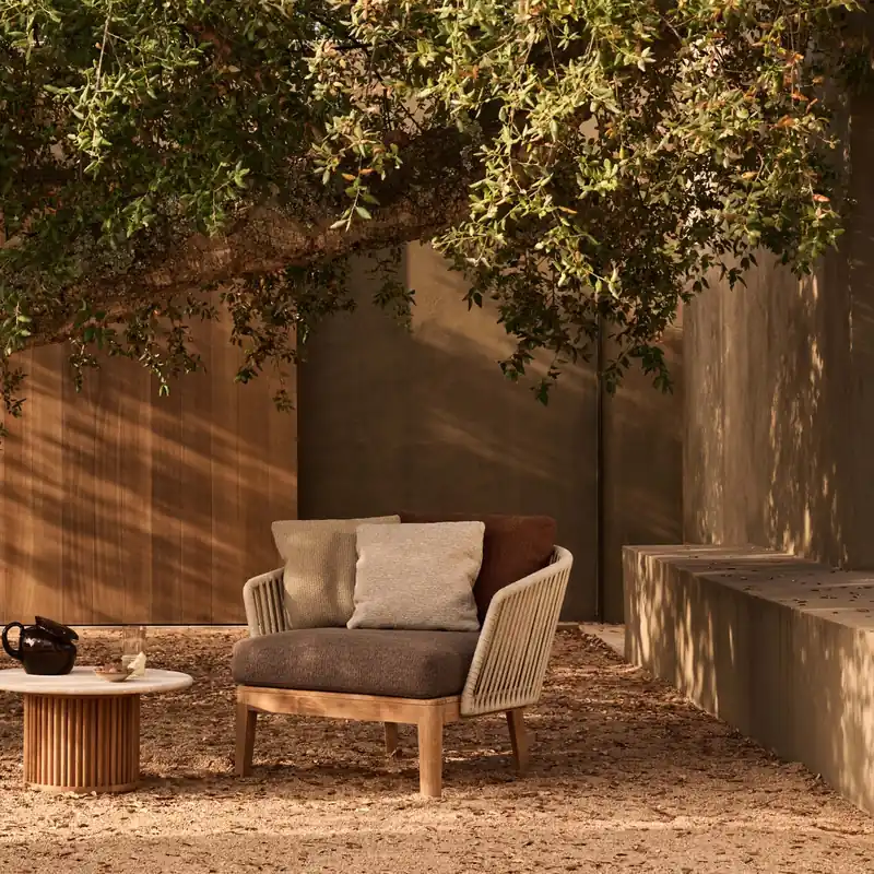 Tribù Mood lounge chair with cream cushions and a small round table on gravel under a leaning tree, a wooden wall behind