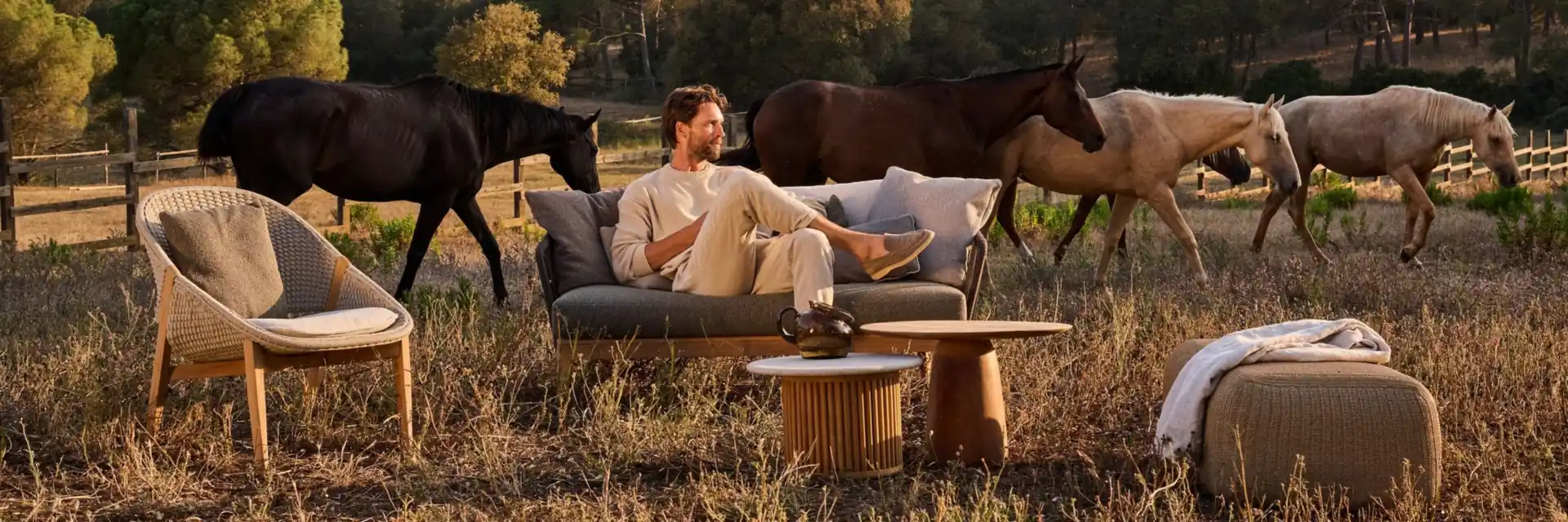 Man lying on a Tribù Mood sofa in a meadow at dusk, with horses grazing behind