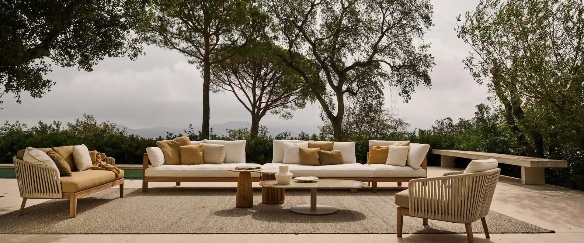 Tribù Mood long sofa with armchairs and low tables on a rug, with trees and a pale sky behind