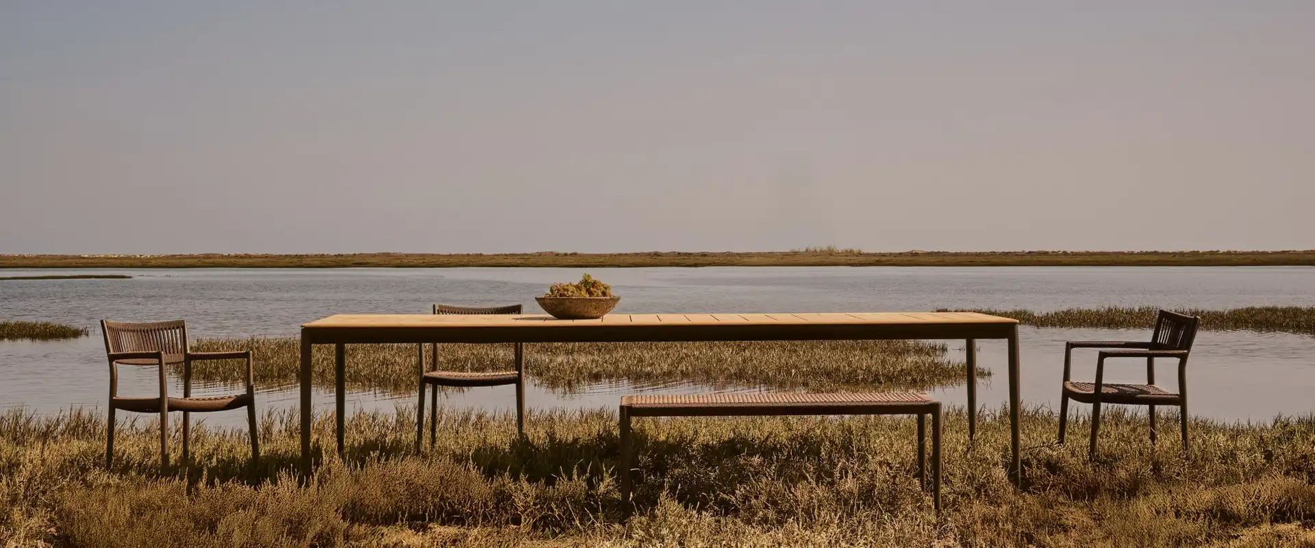 Tribù Nagomi long dining table with chairs and a bench standing in marsh grass at the edge of a calm lagoon