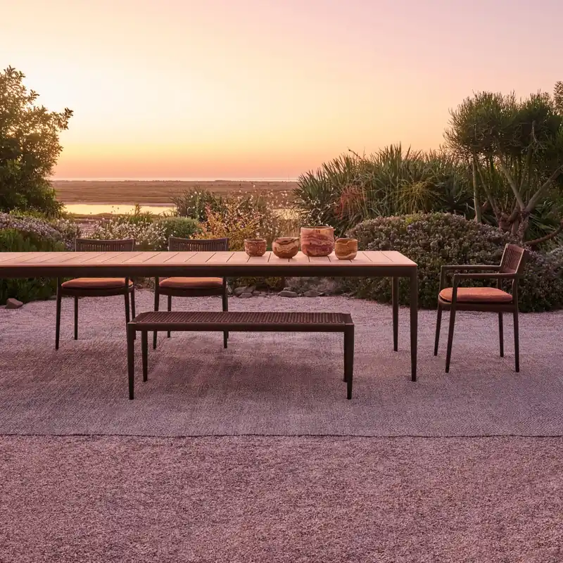 Tribù Nagomi teak dining table with a bench and chairs on gravel under a pink sunset sky, palms and shrubs around