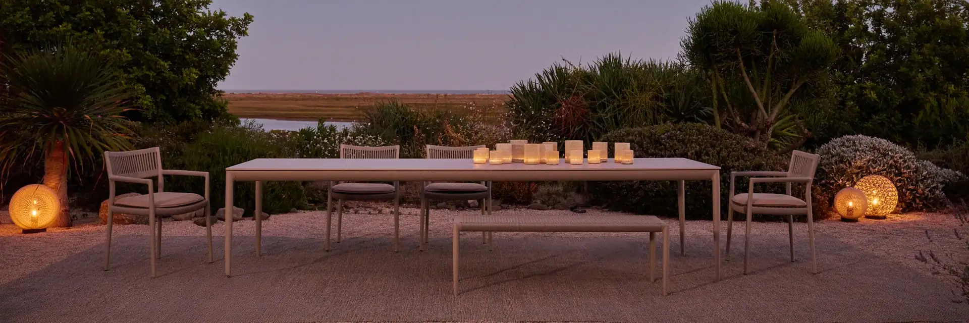 Tribù Nagomi long dining table and chairs on a terrace at dusk, with lanterns and a lagoon behind