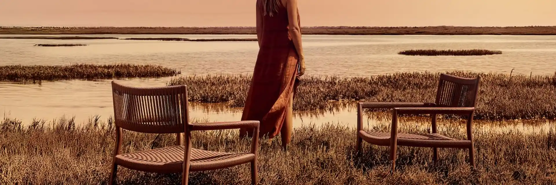 Woman standing between two Tribù Nagomi lounge chairs in the grass beside a lagoon at sunset