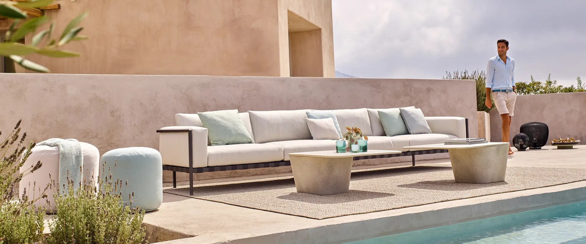 Tribù Natal Alu corner sofa, poufs and a round coffee table on the pool terrace of an adobe-coloured villa, with a man walking past