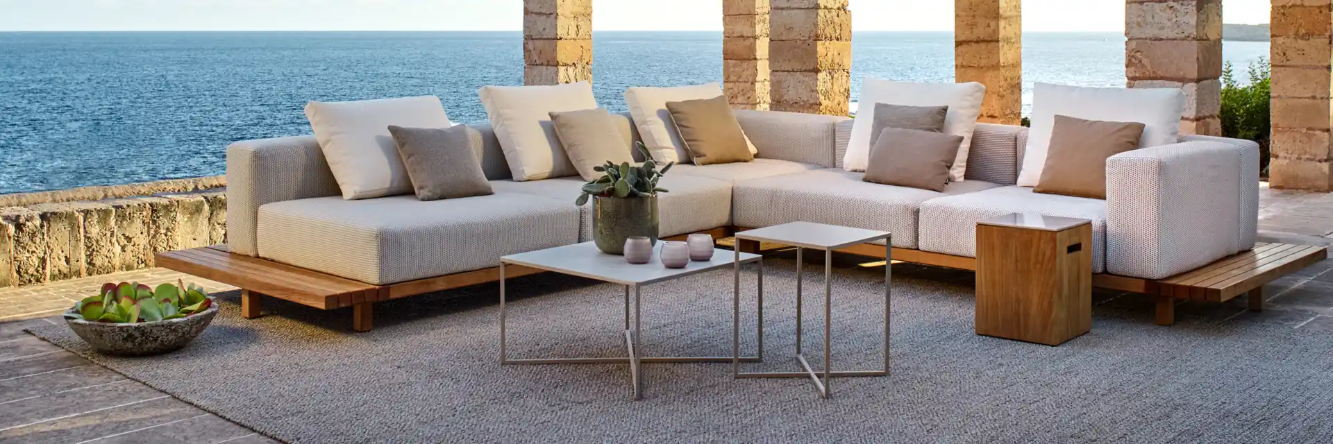 Tribù Natal Alu corner sofa and low tables on a terrace beside a stone pergola, overlooking the sea