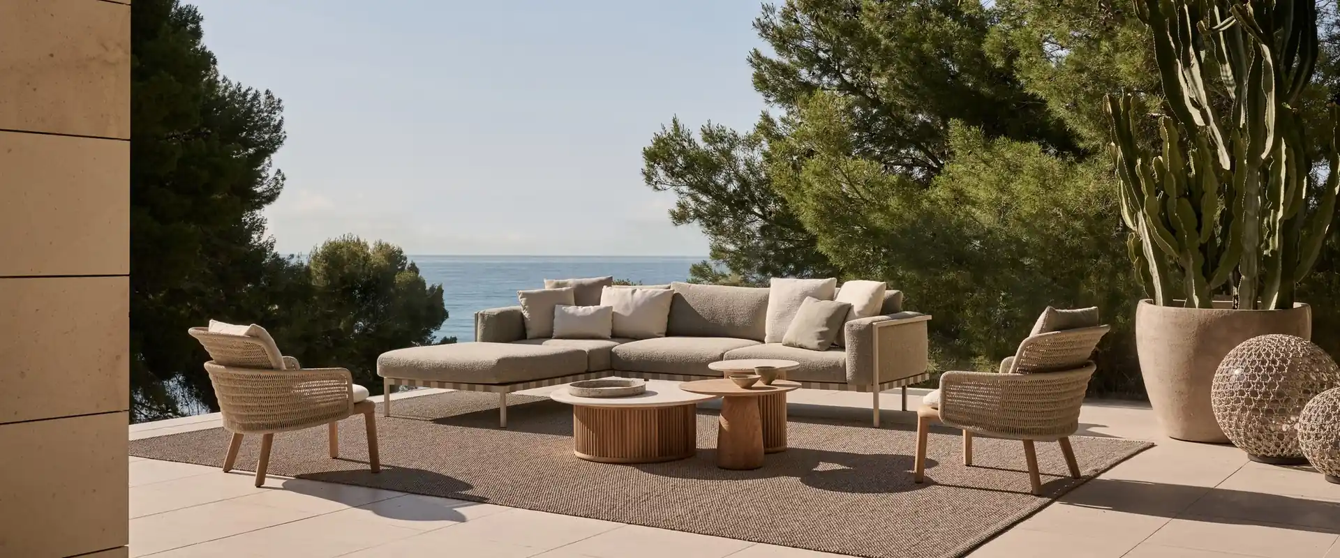 Tribù Natal Alu sofa with lounge chairs on a stone terrace framed by palm fronds and pines, the sea beyond