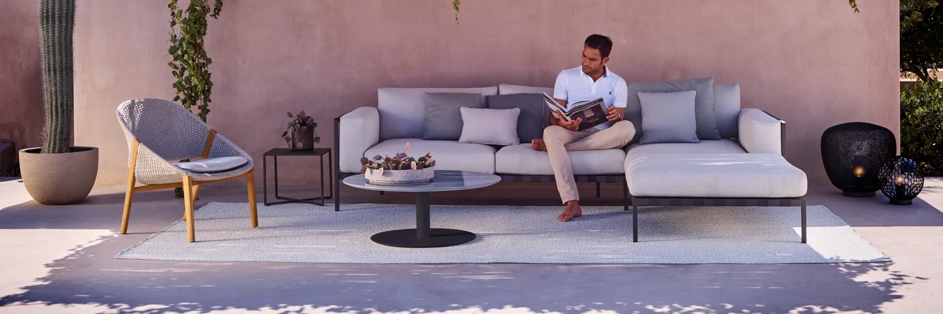 Man reading on a Tribù Natal Alu aluminium sofa in front of a pink rendered wall with a climbing vine, beside an armchair and a cactus