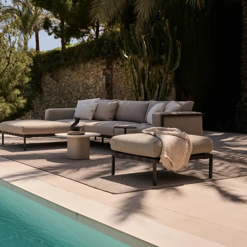 Tribù Natal Alu modular sofa with a chaise on a stone terrace along a turquoise pool, pines behind