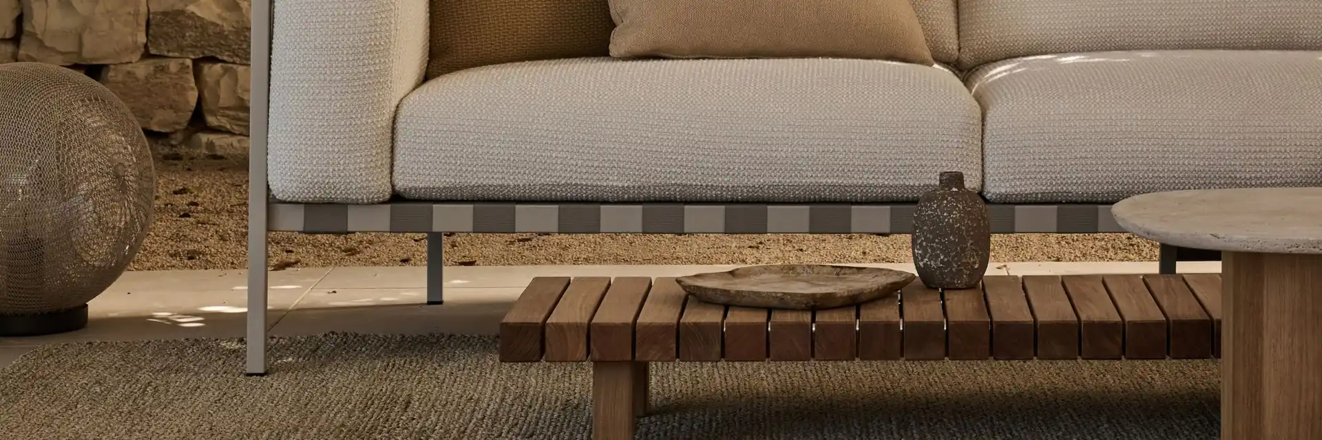 Tribù Natal Alu sofa with cream cushions and a slatted teak coffee table in front of a dry-stone wall