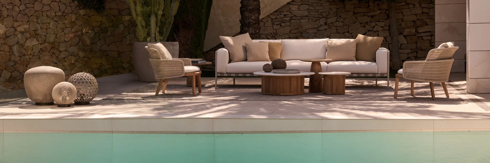Tribù Natal Alu sofa and armchairs on a terrace beside a turquoise pool, with palm trees and a stone wall behind
