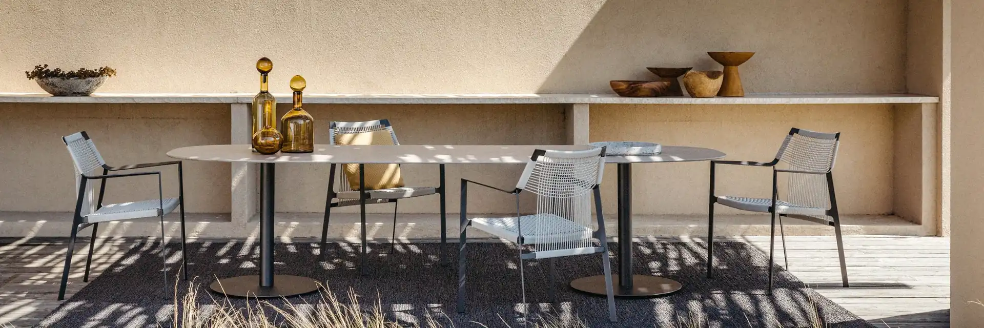 Tribù Nodi dining chairs at a long table on a terrace beside a sand-coloured wall with a built-in shelf