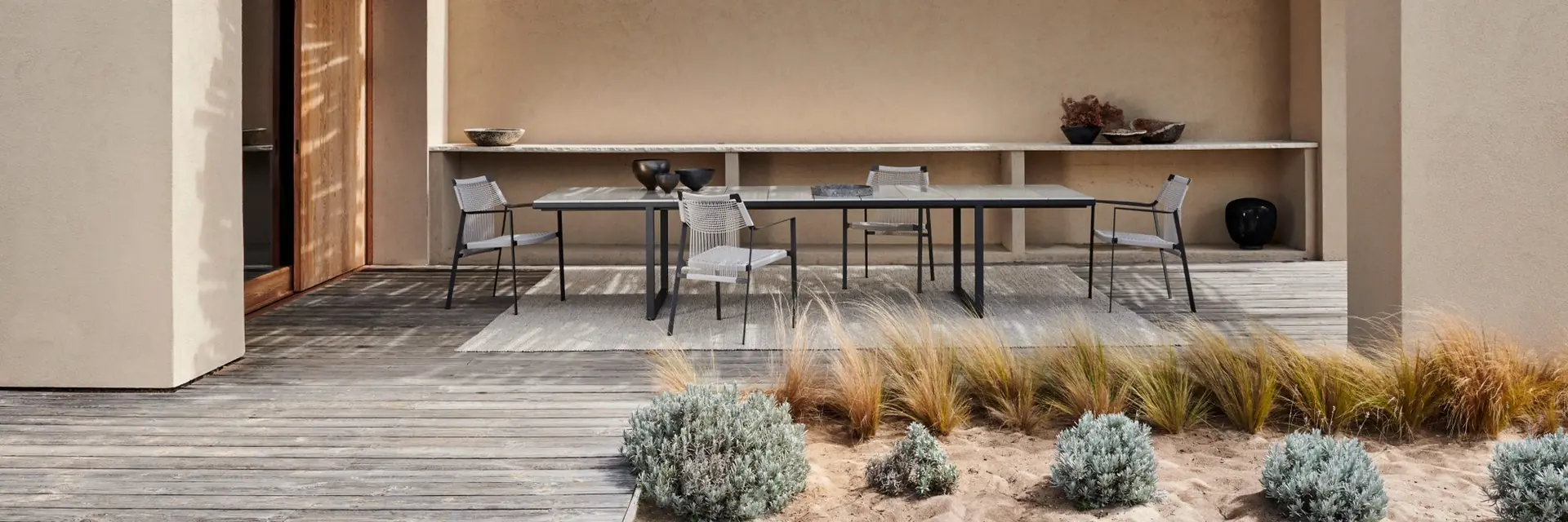 Tribù Nodi dining chairs at a long table on a wooden deck beside a sand-colored house with a dark pergola