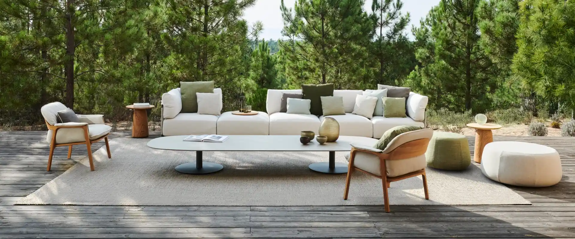 Tribù Nodi sofa with armchairs, poufs and low tables on a wooden deck surrounded by pine trees