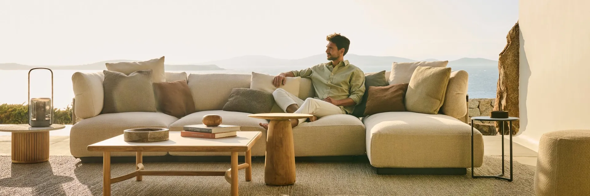 Man sitting on a Tribù Nodi sofa with low tables on a terrace overlooking the sea in soft evening light