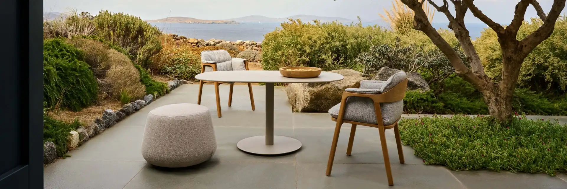 Tribù Nomad teak chair at a round table on a terrace under a tree, with the sea in the distance