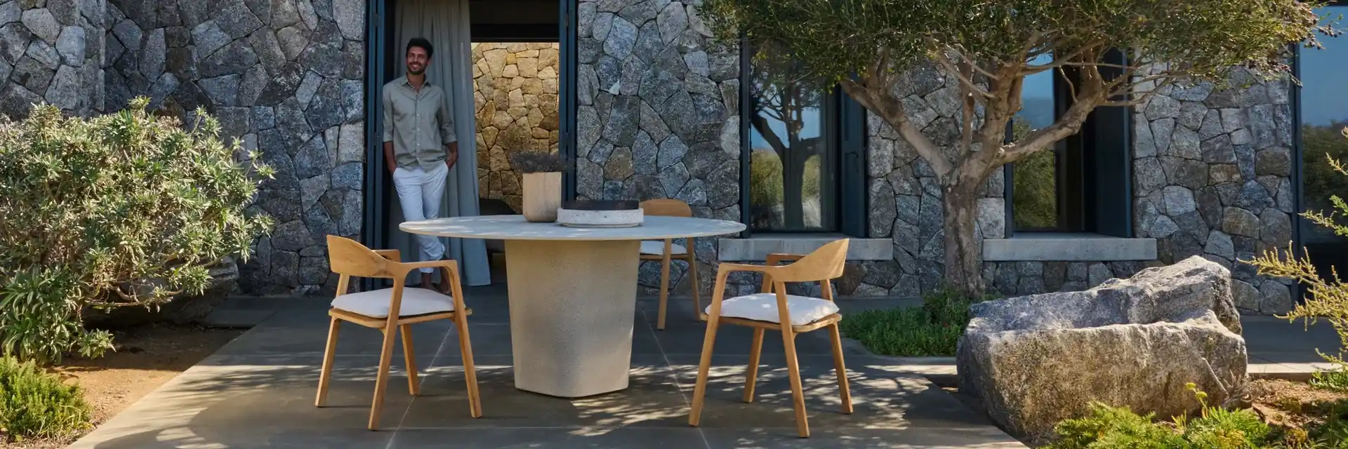 Tribù Nomad teak chairs around a round table on a terrace outside a stone house, beside an olive tree
