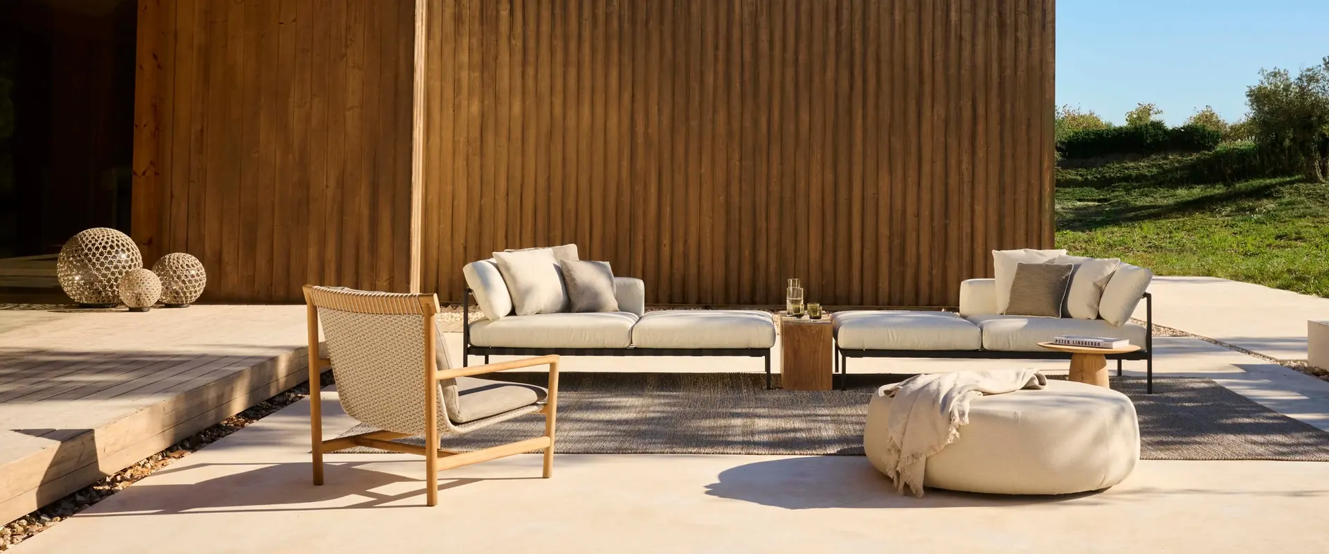 Tribù Nomad low sofa, armchair and poufs on a pale terrace beside a timber-clad pavilion under a blue sky