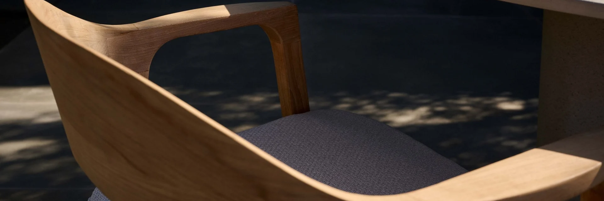 Close-up of the curved teak armrest and back of a Tribù Nomad chair beside a round table, in dappled light