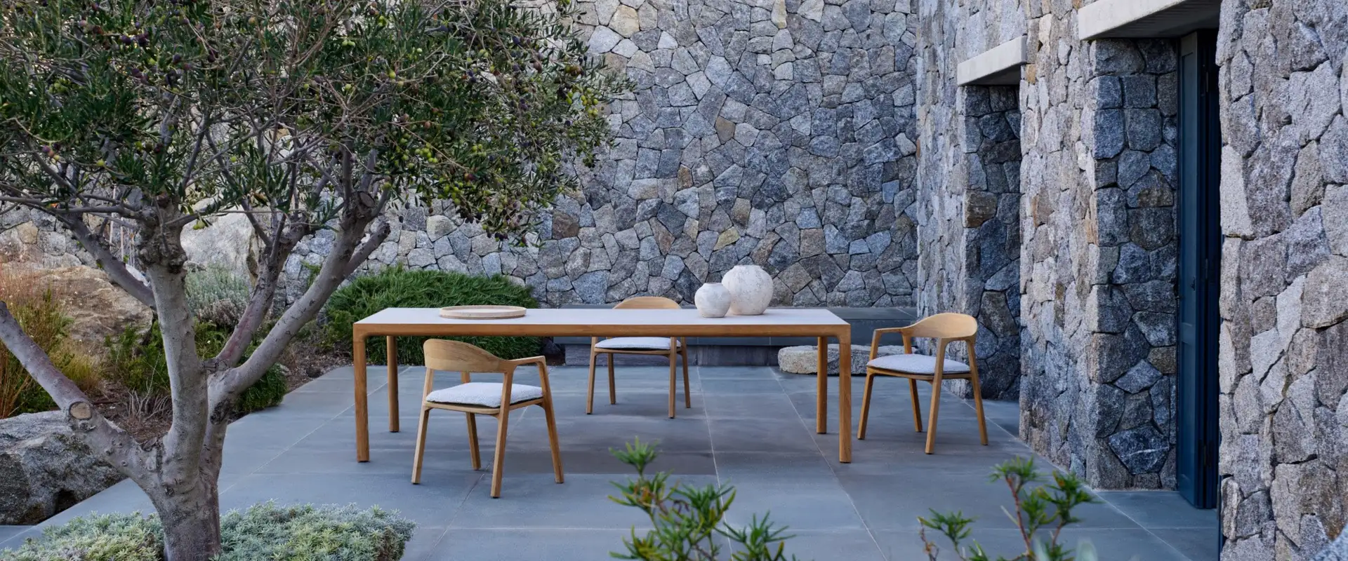 Tribù Nomad teak dining table and chairs on a paved terrace beside a dry-stone house, with an olive tree in the foreground