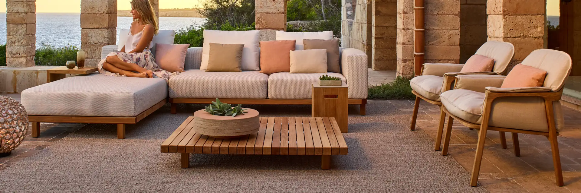 Tribù Nomad teak sofa, easy chairs and coffee table under a stone portico with a tiled roof, with the sea in the distance at sunset