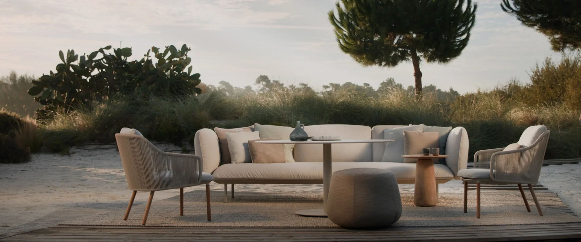 Tribù sofa, lounge chairs and a round pouf on a terrace among dune grasses, with an umbrella pine against a pale sky